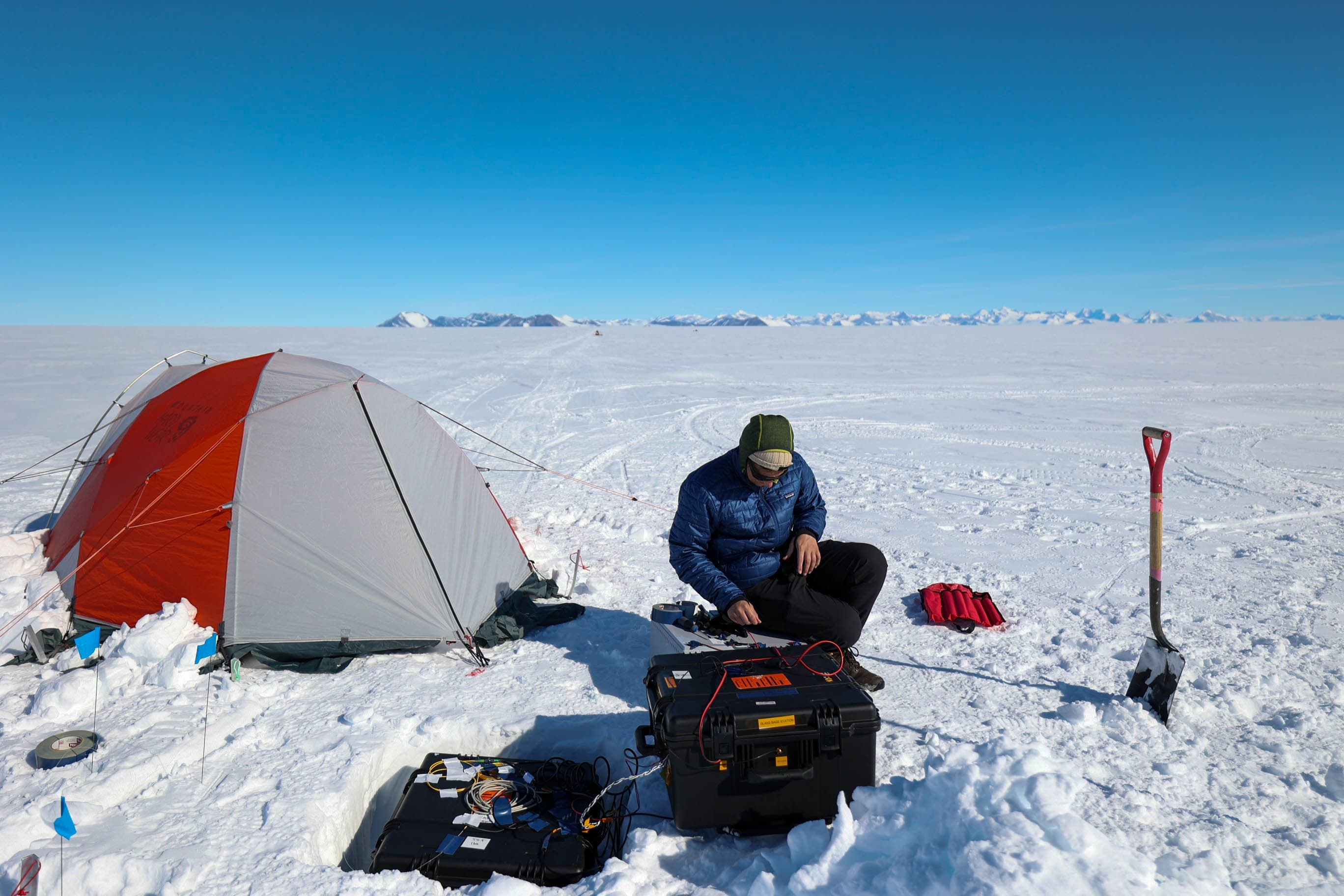 A team member works with equipment cases in the snow beside an orange-and-grey tent.