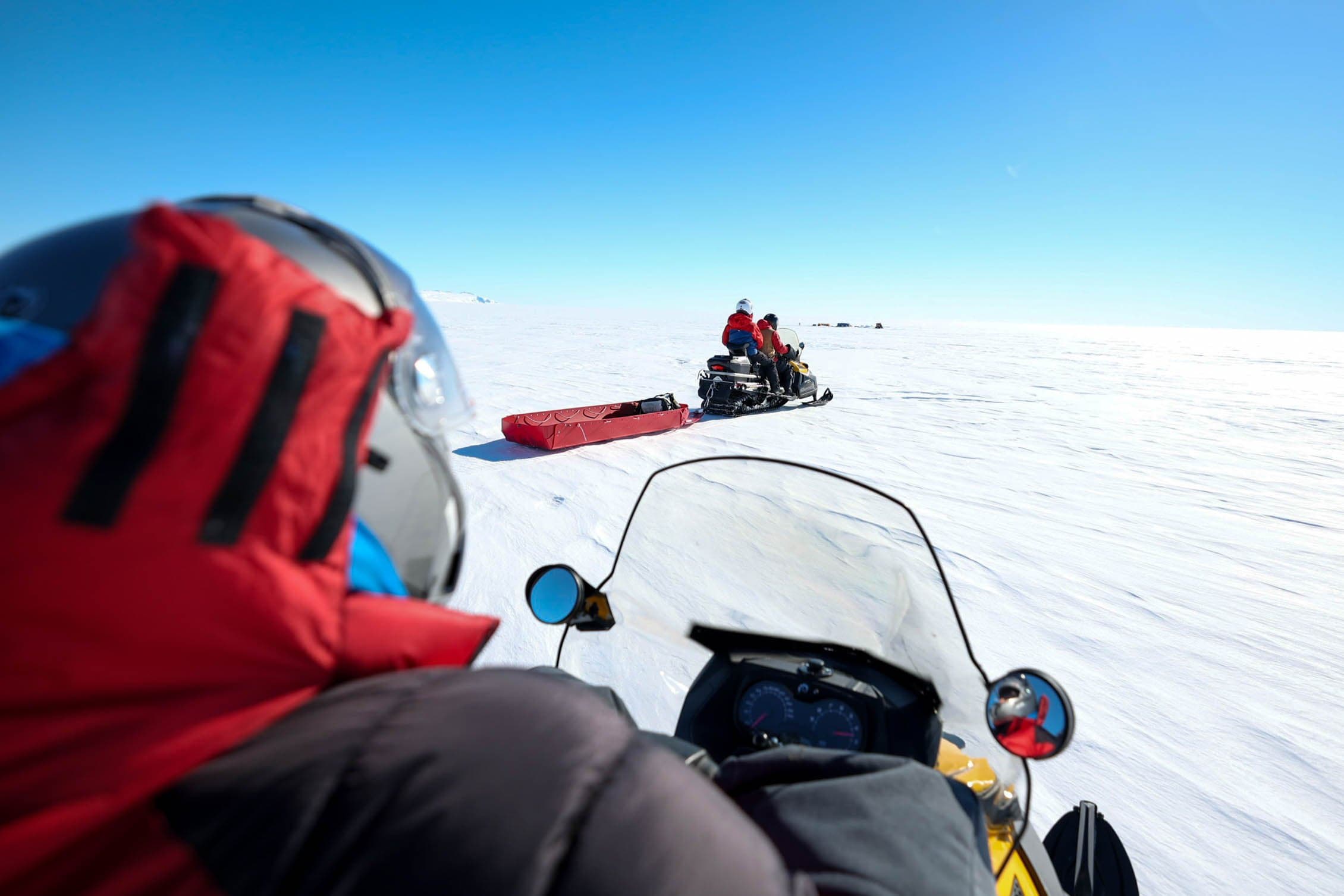 View from a snowmobile following another snowmobile towing a red sled across the ice.