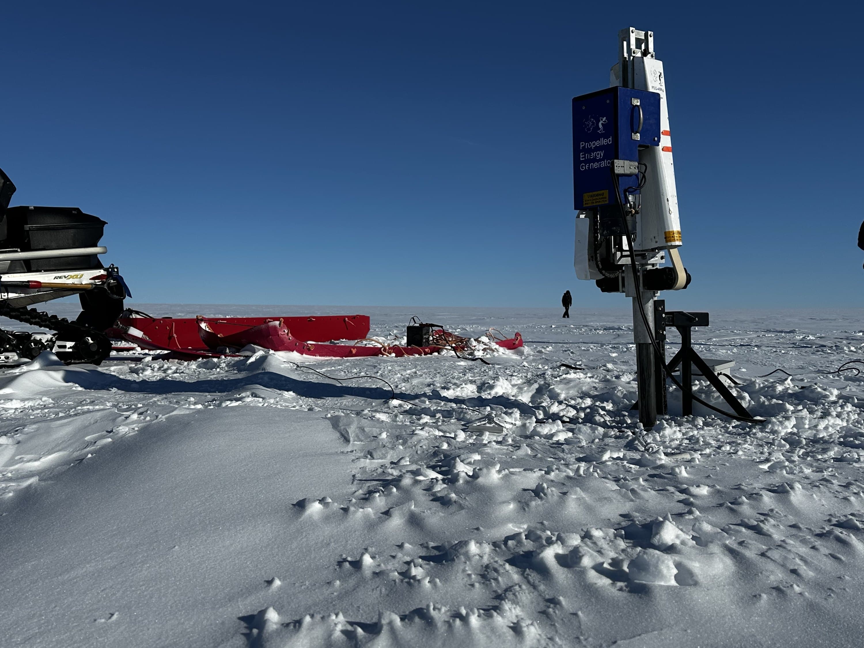 The Propelled Energy Generator, a tall instrument on a tripod, stands on the snow with snowmobiles and sleds behind it.