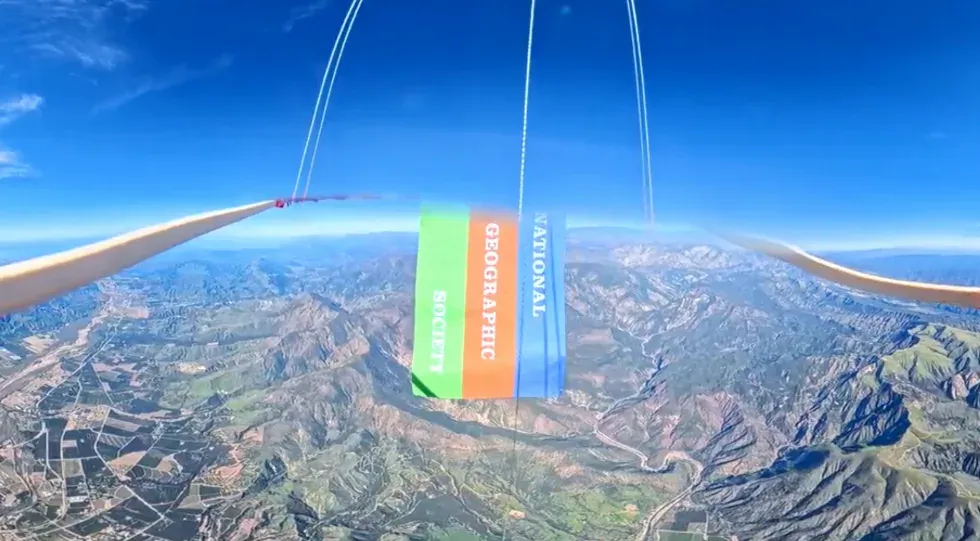 A National Geographic Society banner suspended from a high-altitude balloon above mountainous terrain